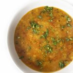 A bowl of yellow arhar dal garnished with chopped fresh cilantro, set against a white background.