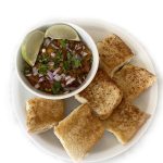 A plate with six pieces of toasted bread, also known as Pav, and a bowl of spicy Instant Pot Pav Bhaji, topped with chopped onions, cilantro, and lime wedges, served on a white background.