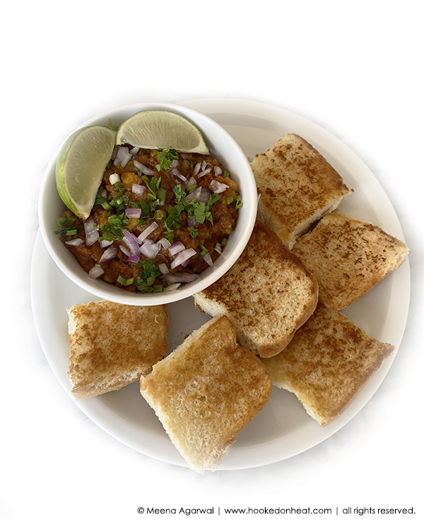 A plate with six pieces of toasted bread, also known as Pav, and a bowl of spicy Instant Pot Pav Bhaji, topped with chopped onions, cilantro, and lime wedges, served on a white background.