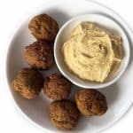 Super Simple Falafels - a Meatless Monday Meal Idea A white plate with six falafel balls and a small white bowl of creamy hummus placed beside them. The plate is set on a white background.