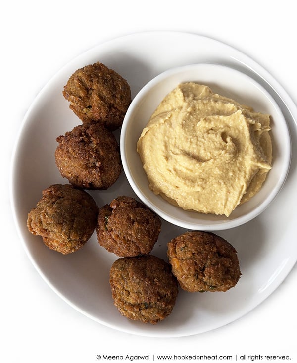 A white plate with six falafel balls and a small white bowl of creamy hummus placed beside them. The plate is set on a white background.